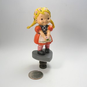 May include: A small, ceramic figurine of a girl with blonde pigtails wearing a red and white dress and holding a flower. The figurine is standing on a gray heart-shaped base.
