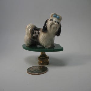 May include: A ceramic Shih Tzu dog figurine with black and white fur and a blue bow, standing on a green glass tabletop. The tabletop is supported by a brass-colored base. A quarter coin is next to the base.