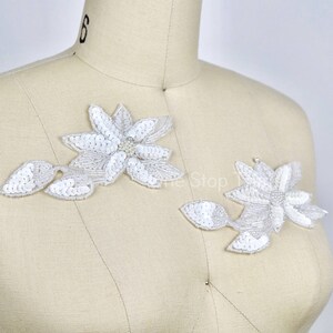 May include: Two white floral appliques with pearl accents. The appliques are on a white fabric background.