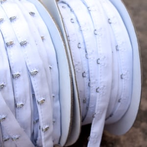 May include: Two spools of white elastic bra strap with metal hooks. The spools are on a white background.
