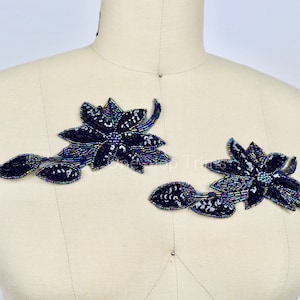 May include: Two iridescent blue floral appliques with a beaded design. The appliques are on a white fabric background.