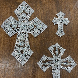 Może przedstawiać: Trzy srebrne krzyże ozdobione kryształkami w różnych rozmiarach. Największy krzyż ma kształt tradycyjnego krzyża z kwadratowym środkiem. Średni krzyż to bardziej ozdobny wzór z zaokrąglonym środkiem. Najmniejszy krzyż to prosty, tradycyjny krzyż.