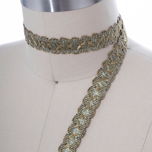 May include: A close-up of a dress form with two pieces of gold and green trim. The trim is a decorative braid with a scalloped edge.