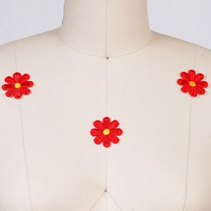 Könnte beinhalten: Drei rote Gänseblümchen-Blumenaufnäher mit gelben Zentren werden auf einer beigen Schneiderpuppe präsentiert. Die Aufnäher sind gestickt und haben ein detailliertes Blütenblattdesign. Die Schneiderpuppe hat eine neutrale Farbe und bietet einen sauberen Hintergrund für die farbenfrohen Aufnäher.