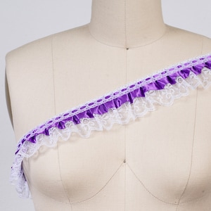 Puede incluir: Cinta de satén morado con ribete de encaje blanco. La cinta está fruncida y tiene un borde festoneado.