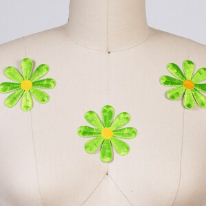 May include: Three green felt daisy flower patches with yellow centers. The patches are on a white fabric background.