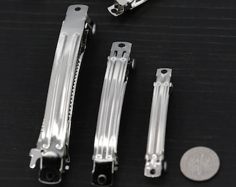May include: A set of silver hair clips in various sizes. The clips are made of metal and have a smooth, shiny finish. The largest clip is about 5 inches long, while the smallest is about 2 inches long.
