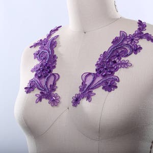 Könnte beinhalten: Lilafarbene Spitzenapplikation mit Perlenbesatz. Die Applikation ist in einem floralen Muster gestaltet und eignet sich perfekt, um jedem Kleidungsstück einen Hauch von Eleganz zu verleihen.
