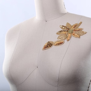 May include: A gold-colored fabric applique in the shape of a flower with a stem. The applique is on a white fabric background.