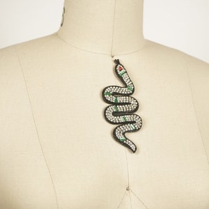 May include: A black snake-shaped pendant with green and white details. The snake is covered in rhinestones and has a red eye.