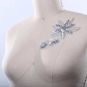 May include: A silver and white beaded flower applique with a pearl center. The applique is attached to a white fabric mannequin.