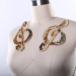 Puede incluir: Dos apliques de clave de sol de lentejuelas doradas sobre un fondo de tela blanca. Los apliques están hechos de lentejuelas doradas y plateadas y son perfectos para agregar un toque de brillo a cualquier proyecto.