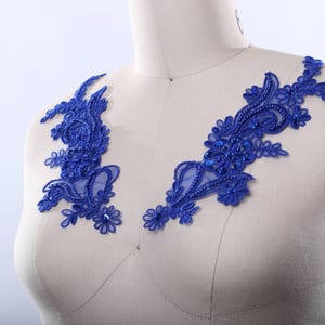 May include: Two royal blue lace appliques with floral and beaded designs are displayed on a mannequin. The appliques are symmetrical, with intricate patterns and a delicate, airy appearance, perfect for embellishing garments.