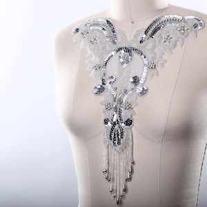 May include: A silver and white beaded and sequined lace applique with a dangling fringe. The applique is shaped like a V and has a floral design.