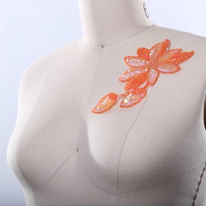 May include: Orange beaded flower applique with a pearl-like center. The applique is on a white fabric background.