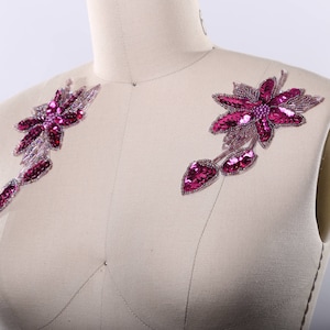 May include: Two pink floral appliques with sequins and beads. The appliques are on a white fabric background.