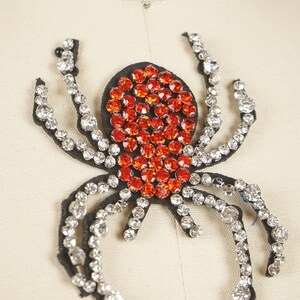 May include: A black spider applique with a body of orange rhinestones and legs outlined with clear rhinestones. This decorative embellishment is ideal for adding sparkle to clothing or accessories.