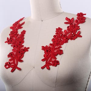 May include: Two red lace appliques with floral designs and sequins. The appliques are on a white fabric background.
