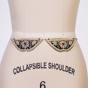May include: Two black fabric embellishments with gold embroidery and colorful jewels. The embellishments are shaped like a crescent moon and are attached to a white fabric belt.