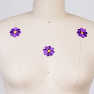 May include: Three purple fabric flower patches with yellow centers, sewn onto a white fabric mannequin torso.