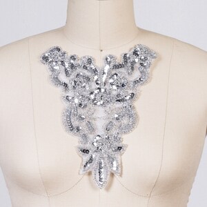 May include: Silver beaded applique with a floral design. The applique is made of sequins and beads, and is attached to a white mesh backing. The applique is displayed on a mannequin.