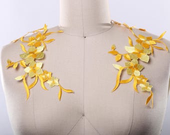 May include: Two yellow floral appliques with delicate flower and leaf designs. The appliques are on a light brown fabric background.