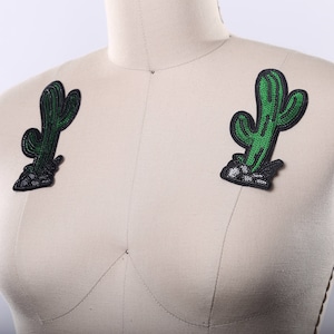 May include: Two green cactus iron-on patches with black outlines and white stitching. The patches are on a white fabric background.