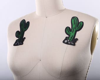 May include: Two green cactus iron-on patches with black outlines and white stitching. The patches are on a white fabric background.