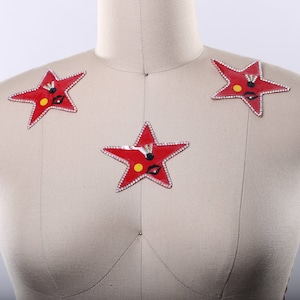 May include: Three red star-shaped patches with rhinestones and a black and yellow design. The patches are on a beige fabric background.