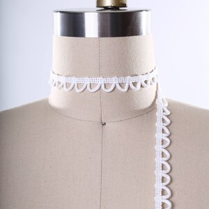 May include: White scalloped trim lace on a dress form. The lace is shown in a continuous length, with a portion of it wrapped around the neck of the form.