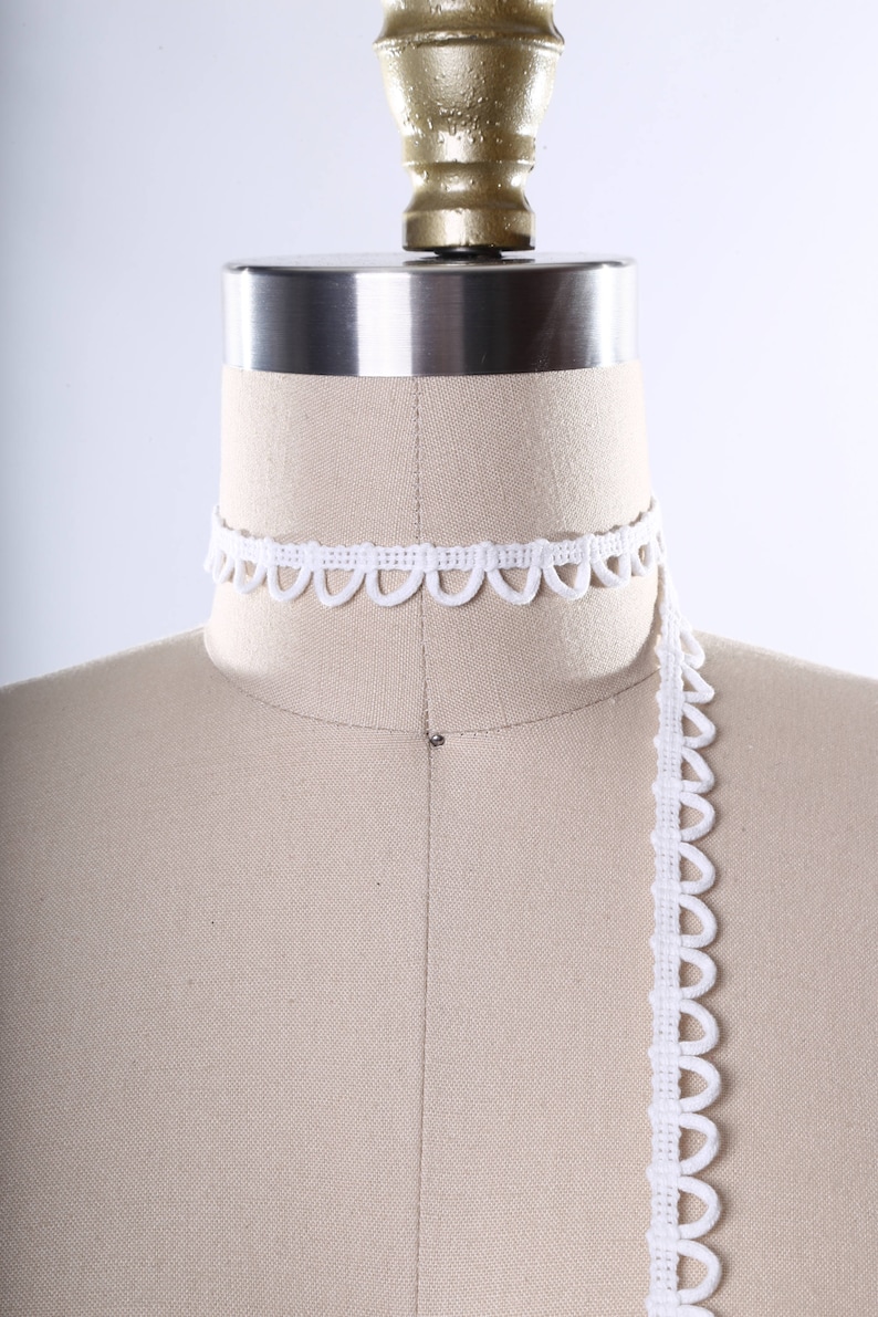 May include: White scalloped trim lace on a dress form. The lace is shown in a continuous length, with a portion of it wrapped around the neck of the form.
