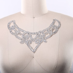 May include: A silver rhinestone embellishment in the shape of a curved, open-ended collar. The embellishment is made of small, clear rhinestones that are sewn together in a delicate pattern.