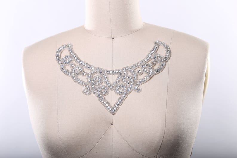 May include: A silver rhinestone embellishment in the shape of a curved, open-ended collar. The embellishment is made of small, clear rhinestones that are sewn together in a delicate pattern.
