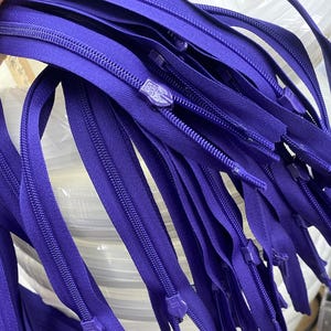 May include: A bundle of purple zipper tape with metal zipper pulls. The tape is arranged in a stack, with the zipper pulls visible at the bottom.