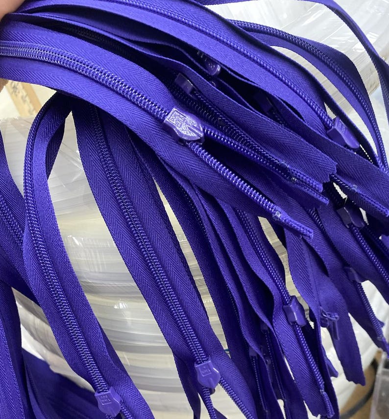May include: A bundle of purple zipper tape with metal zipper pulls. The tape is arranged in a stack, with the zipper pulls visible at the bottom.