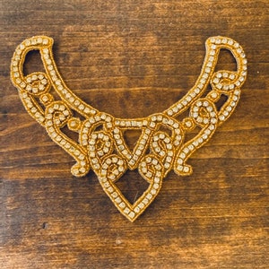 Könnte beinhalten: Ein goldfarbener, mit Strass besetzter, dekorativer Besatz mit einem gewellten Design.