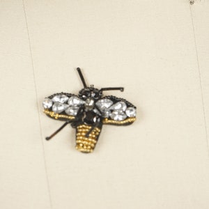 May include: A black and gold bee-shaped brooch with clear rhinestones. The brooch is made of beads and is a decorative accessory.