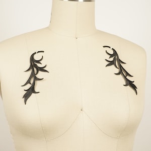 Puede incluir: Dos apliques de tela negra en forma de hojas estilizadas, fijados a un maniquí de tela blanco.