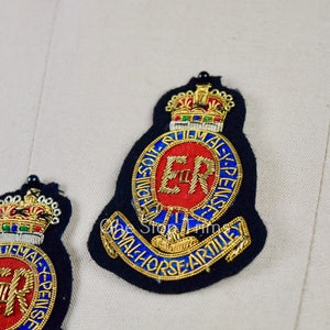 May include: Two embroidered patches with a gold crown, a red circle with the letters "ER" in gold, and the text "ROYAL HORSE ARTILLERY" in gold. The patches are on a white fabric background.
