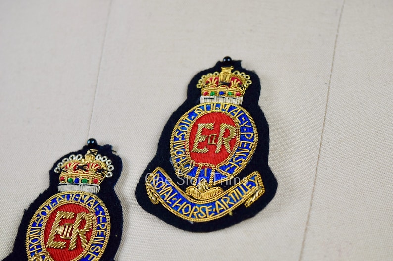 May include: Two embroidered patches with a gold crown, a red circle with the letters "ER" in gold, and the text "ROYAL HORSE ARTILLERY" in gold. The patches are on a white fabric background.