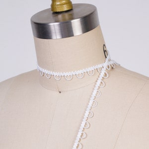 May include: White elastic trim with a scalloped edge. The trim is shown on a dress form.