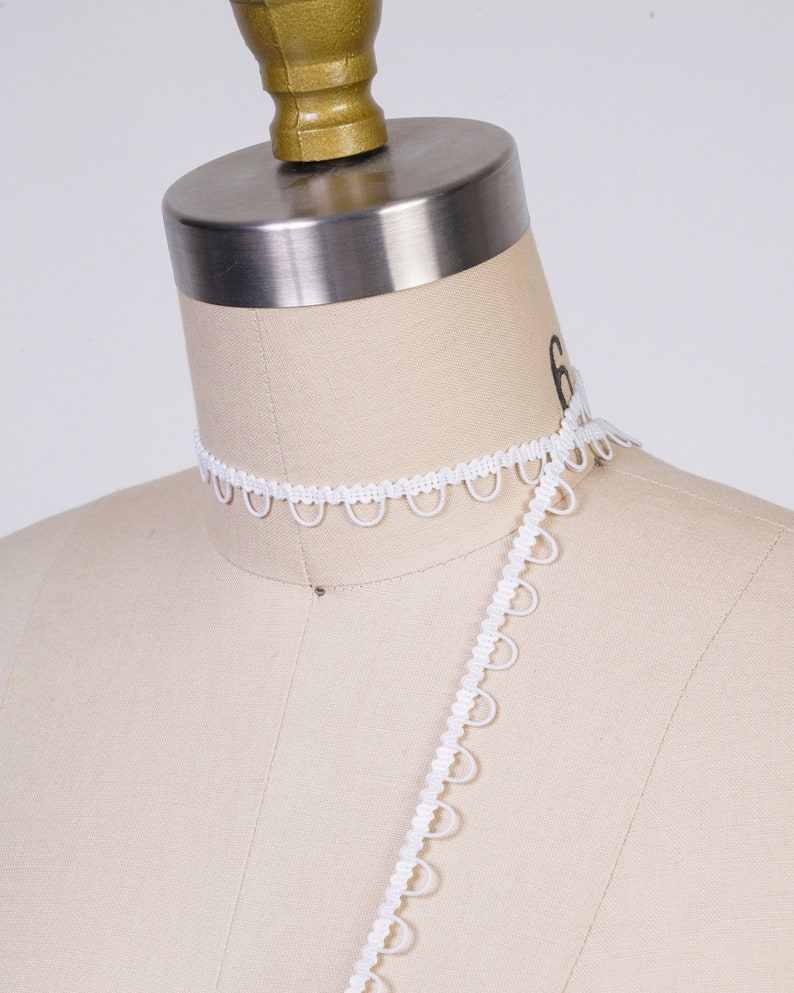 May include: White elastic trim with a scalloped edge. The trim is shown on a dress form.