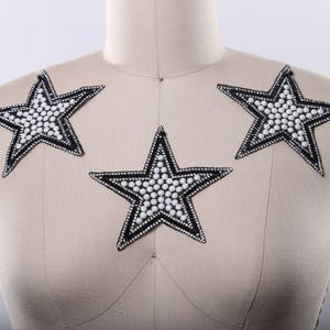 May include: Three star-shaped appliques. Each star has a black outline with white bead and rhinestone embellishments. The stars are arranged on a mannequin, suggesting they are for clothing decoration.