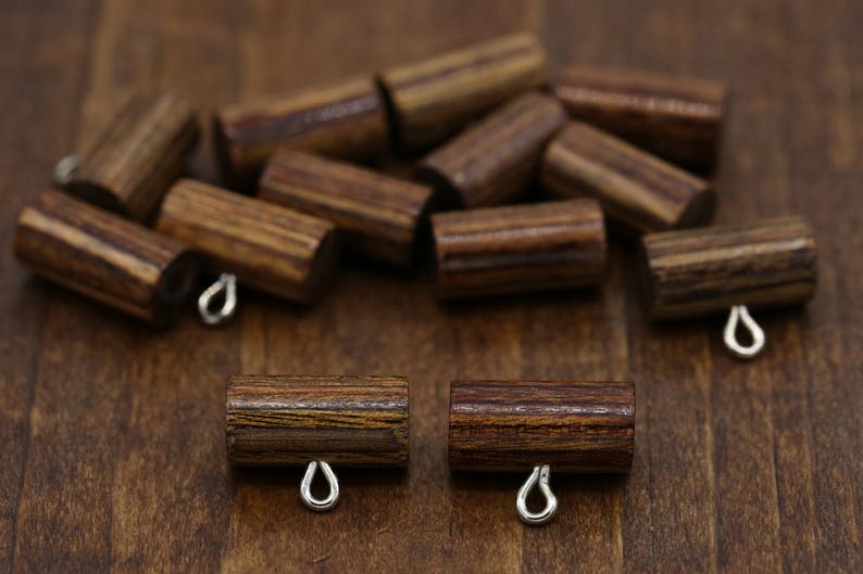 K&ouml;nnte beinhalten: Eine Gruppe von Holzperlen mit einem gestreiften Muster und silbernen Metallschlaufen. Die Perlen sind auf einer Holzoberfl&auml;che zu einem Haufen angeordnet.