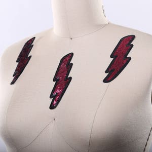 Puede incluir: Tres parches en forma de rayo rojo y negro sobre un fondo de tela blanca. Los parches están hechos de lentejuelas.