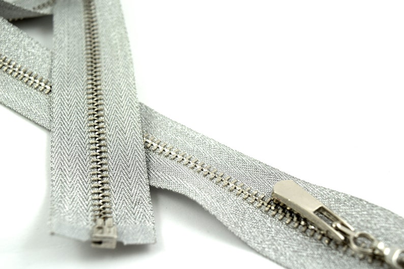 May include: A silver zipper with a silver slider. The zipper is closed and the slider is at the top of the zipper.
