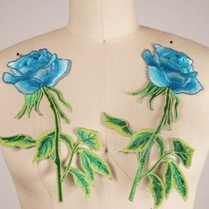 Puede incluir: Dos parches bordados de rosas azules con hojas y tallos verdes. Las rosas son de un azul claro con un contorno azul más oscuro. Las hojas son de un verde brillante con un contorno amarillo.