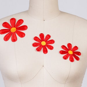 May include: Three red felt flower appliques with yellow centers. The flowers are arranged on a white fabric background.