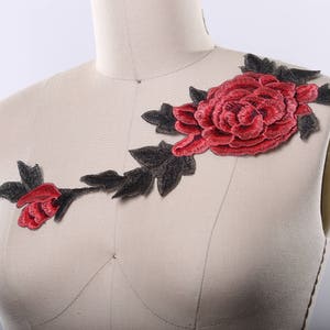 Op de afbeelding: Rode geborduurde bloemenapplicatie met zwarte stelen en bladeren. De applicatie is op een witte stoffen achtergrond.