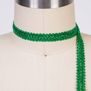 May include: Green metallic trim with a decorative woven pattern. The trim is shown wrapped around a dress form.